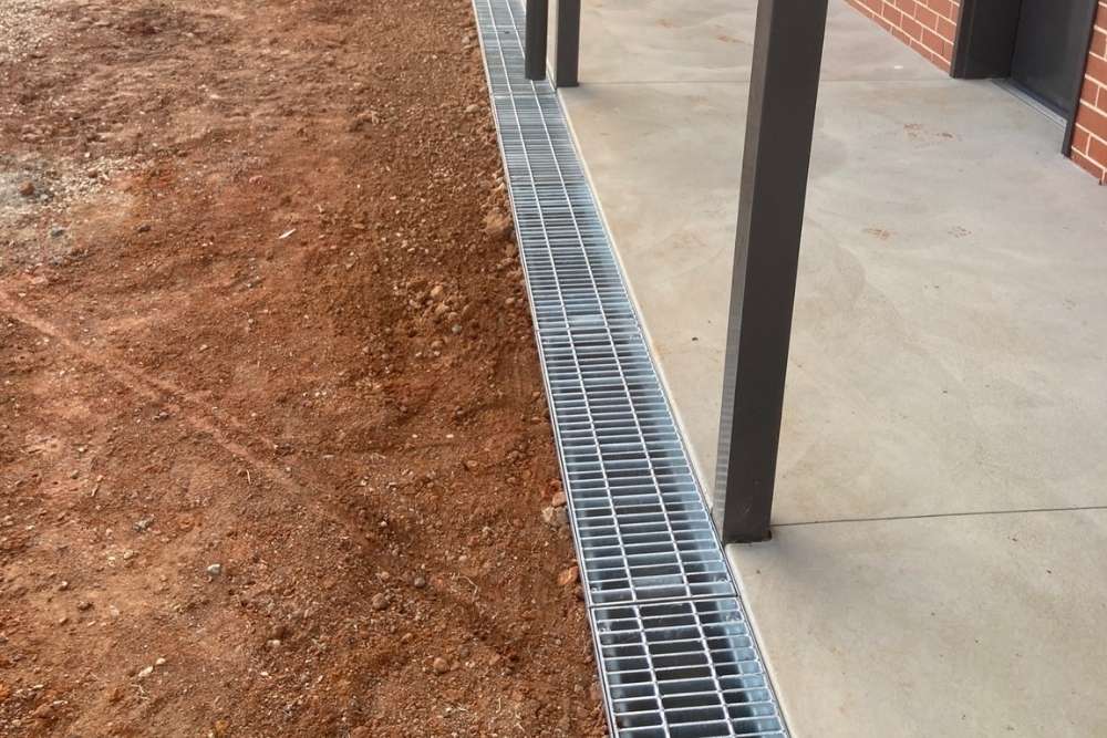 A newly installed silver metal grate drain running along the edge of a concrete patio next to red soil-